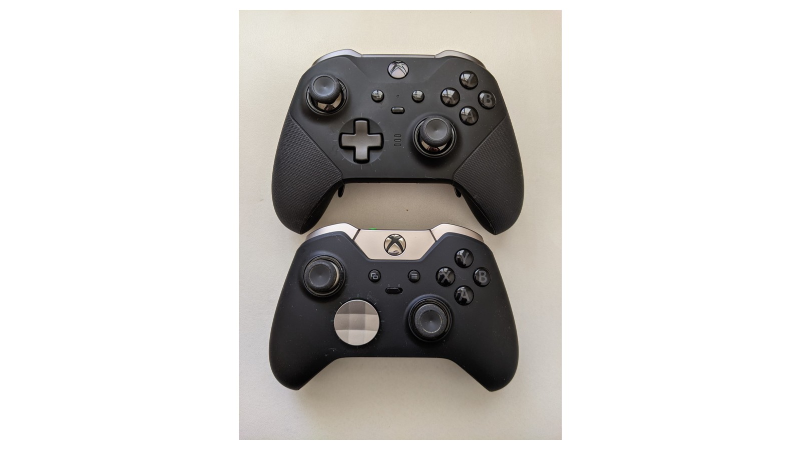 Two black Xbox Elite controllers, with the Series 2 above the original, on a pale surface.