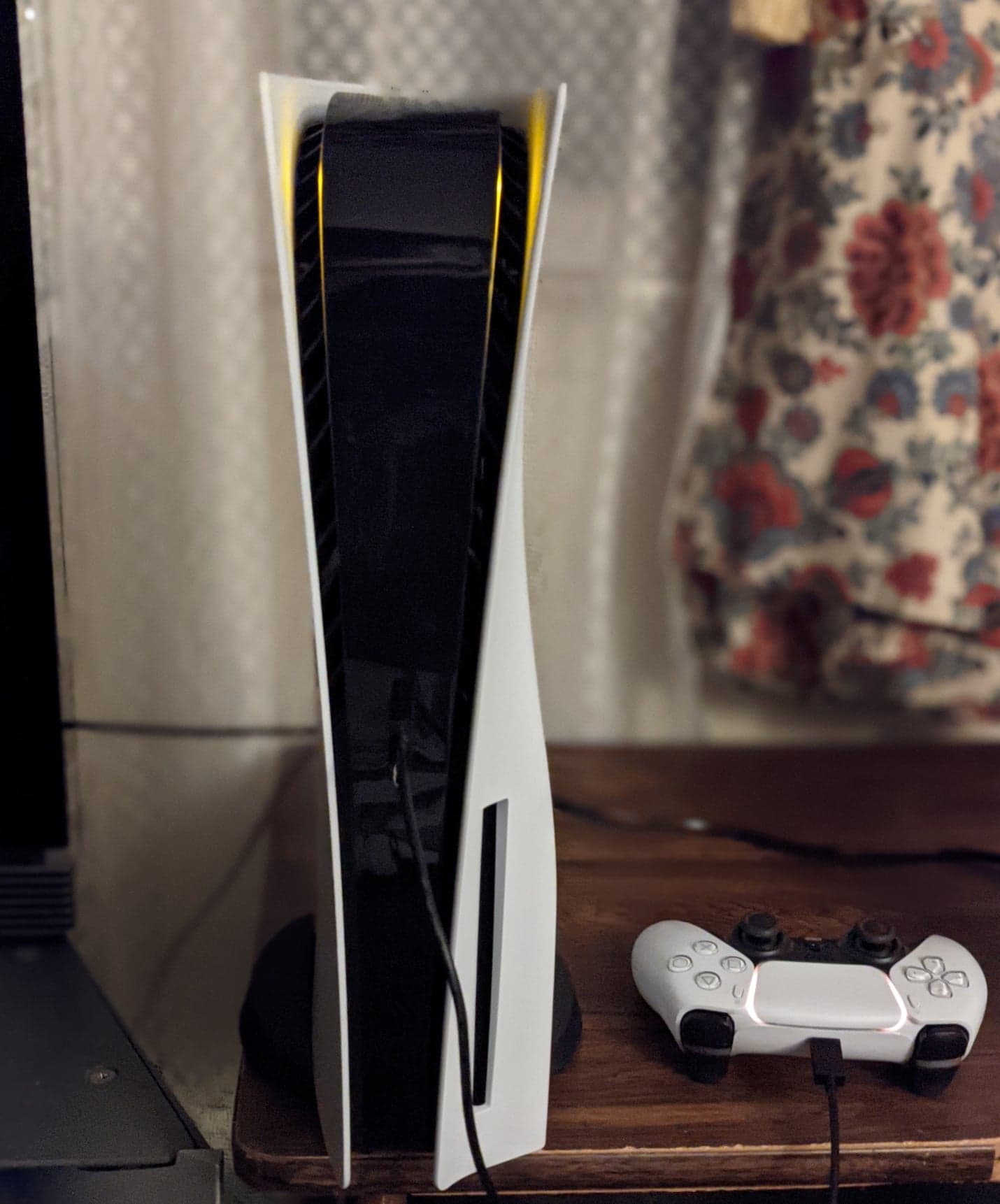 A white PlayStation 5 stands vertically beside a wired DualSense controller on a wooden surface