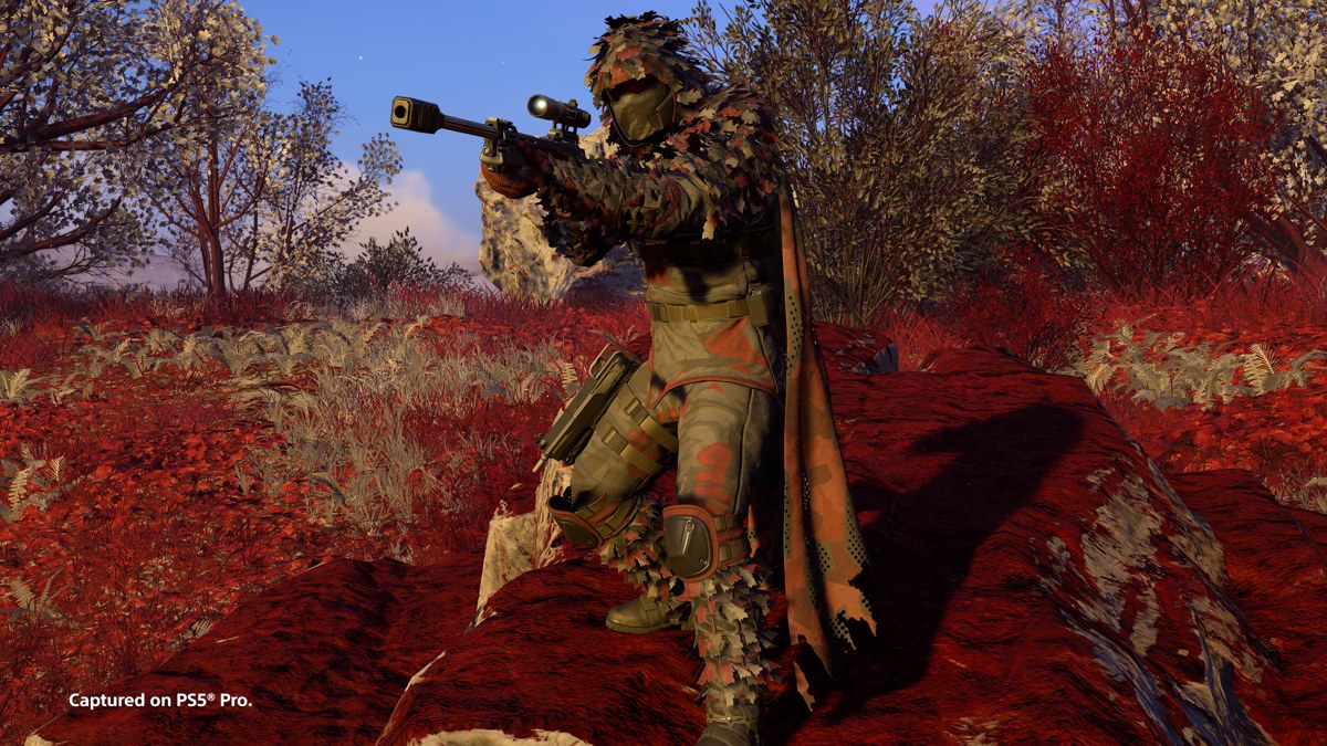 A camouflaged Helldiver aims a scoped rifle among autumn trees in an official Helldivers 2 screenshot.