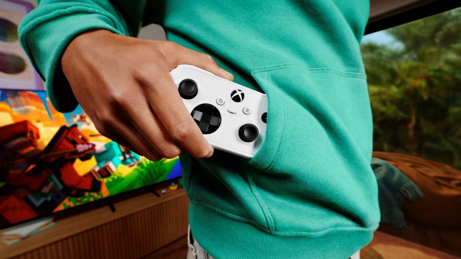 A person in a teal hoodie slides a small white Xbox Nano Controller into the front pocket, with a Minecraft-style game on a TV behind them