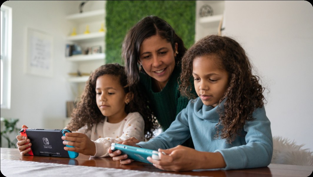 A parent and two children playing Nintendo Switch consoles together at a table